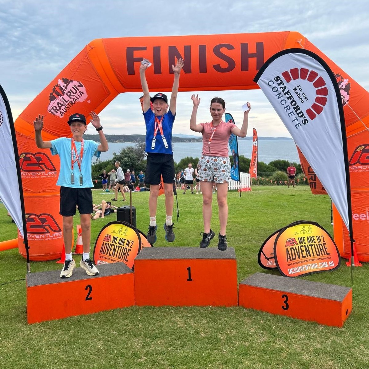 Three young athletes celebrating on the winners’ podium under a Stafford’s Concrete sponsored finish line at the Tathra MultiSport Festival.